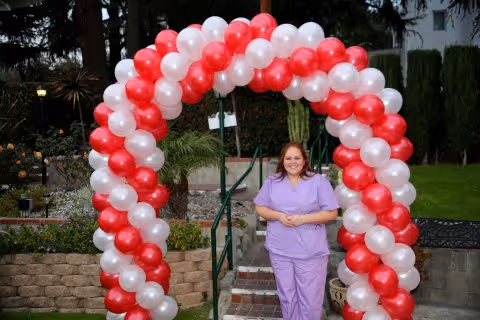 A woman in purple scrubs standing outdoors in front of a red and white balloon arch with greenery and garden elements in the background.