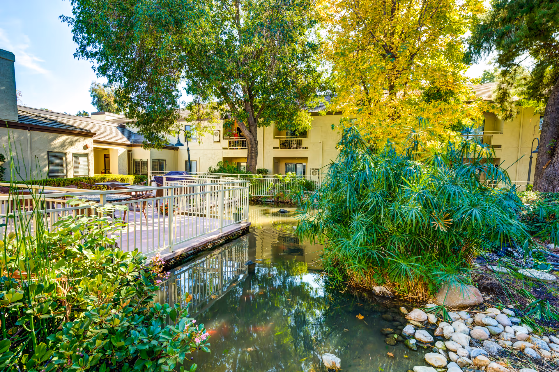 A serene outdoor courtyard at The Oaks at Inglewood featuring a small pond surrounded by lush green plants and trees. There is a patio area with tables and chairs enclosed by a white railing, and the building with balconies and windows is visible in the background under a bright blue sky.