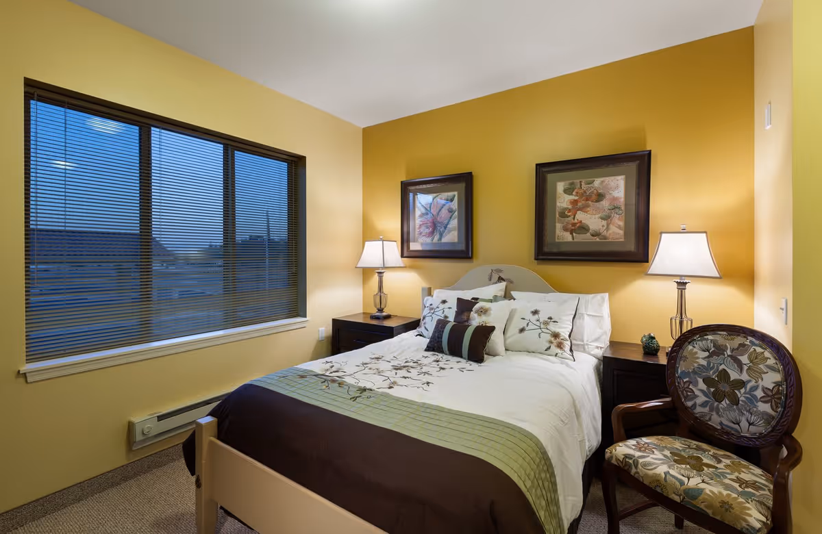 A cozy bedroom with yellow walls featuring a bed with floral-patterned bedding and multiple pillows. On either side of the bed are dark wooden nightstands with lamps. Above the bed are two framed floral artworks. A floral upholstered chair is placed next to the right nightstand. A large window with blinds is on the left wall.
