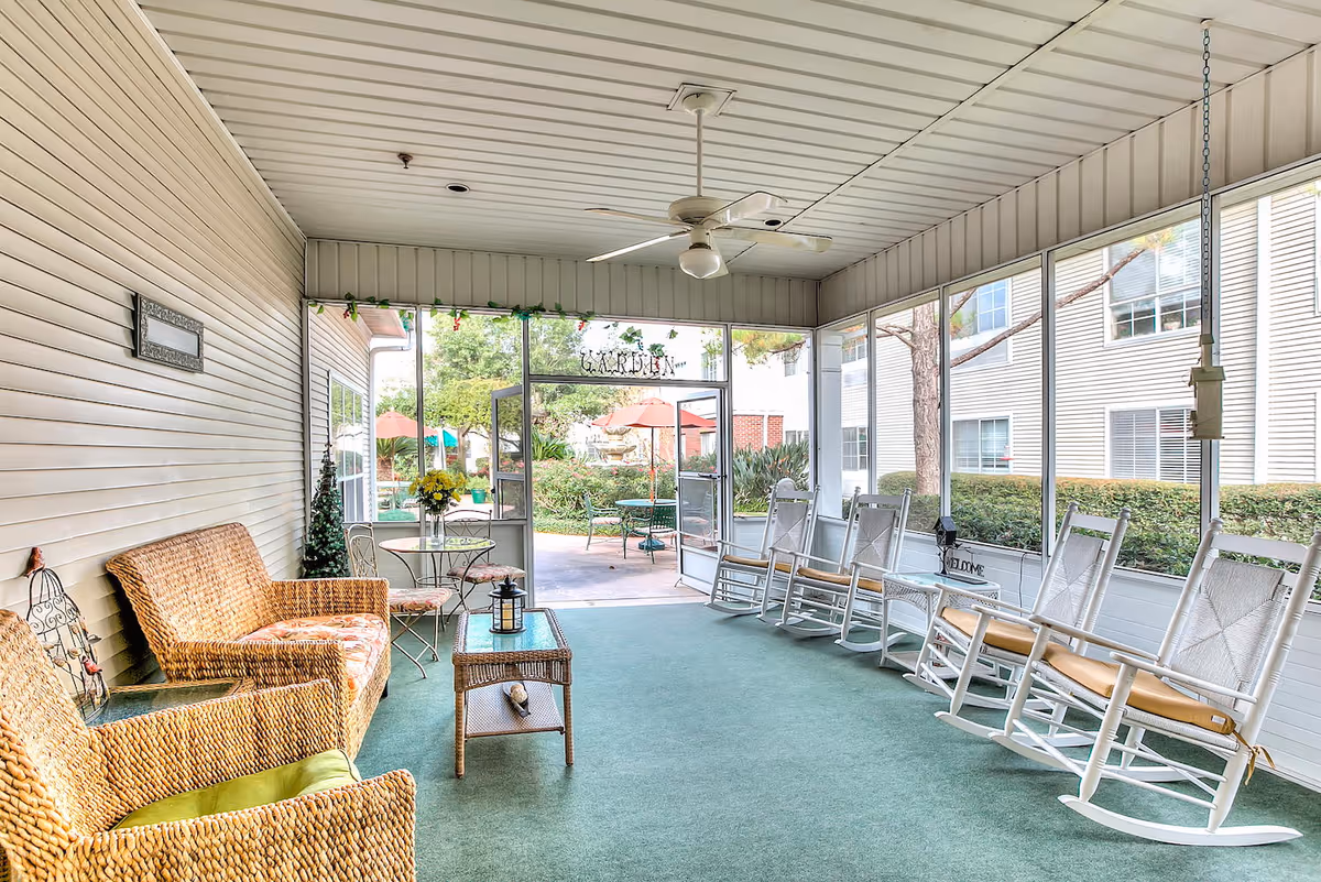 A bright enclosed porch area with wicker furniture including a loveseat and chair, a small glass-top table with a lantern, and a row of white rocking chairs along the right side. The porch has large screened windows providing a view of an outdoor garden area with tables and umbrellas. The ceiling has a white fan and the floor is covered with green carpet.