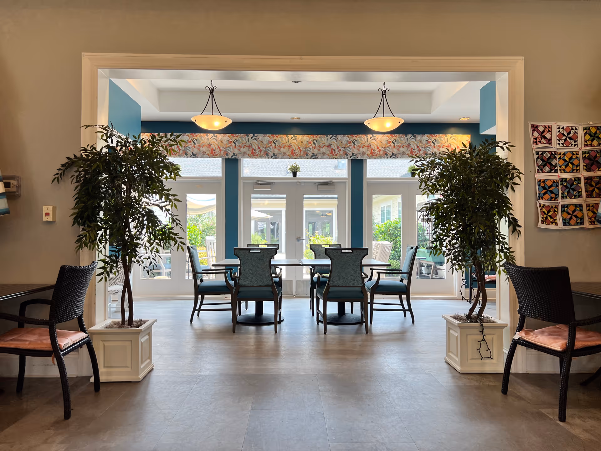 A bright dining area with a table and six chairs arranged around it. The room has large glass doors and windows letting in natural light, with a floral valance above the doors. Two potted plants flank the entrance to the dining area, and two hanging light fixtures illuminate the space. There are additional chairs with cushions on either side of the entrance, and a colorful quilt hangs on the wall to the right.