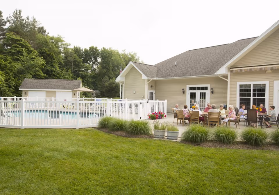 Fenced outdoor pool and patio beside a beige townhome-style building, with a group of people seated at tables on the patio.