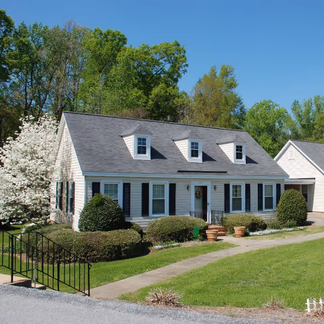 A white single-story house with a gray roof and three dormer windows. The house has black shutters on the windows and a small front porch with a wreath on the door. There are neatly trimmed bushes and a flowering tree in the front yard, with a paved walkway leading to the entrance. The sky is clear and blue, and there are tall green trees in the background.