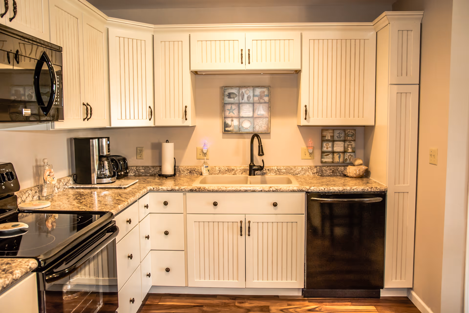 A modern kitchen with white cabinets and drawers, granite countertops, a black electric stove, a black microwave, a coffee maker, a toaster, a paper towel holder, and a black dishwasher. There is a double sink with a black faucet in the center, and decorative wall art above the sink. The floor is wooden.
