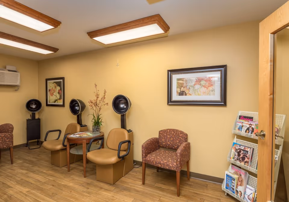 Interior view of a hair salon area in a senior living facility with three vintage hair dryers, two tan salon chairs, a small round table with magazines and a plant, a patterned armchair, framed floral artwork on the walls, and a magazine rack filled with various magazines near a wooden door.