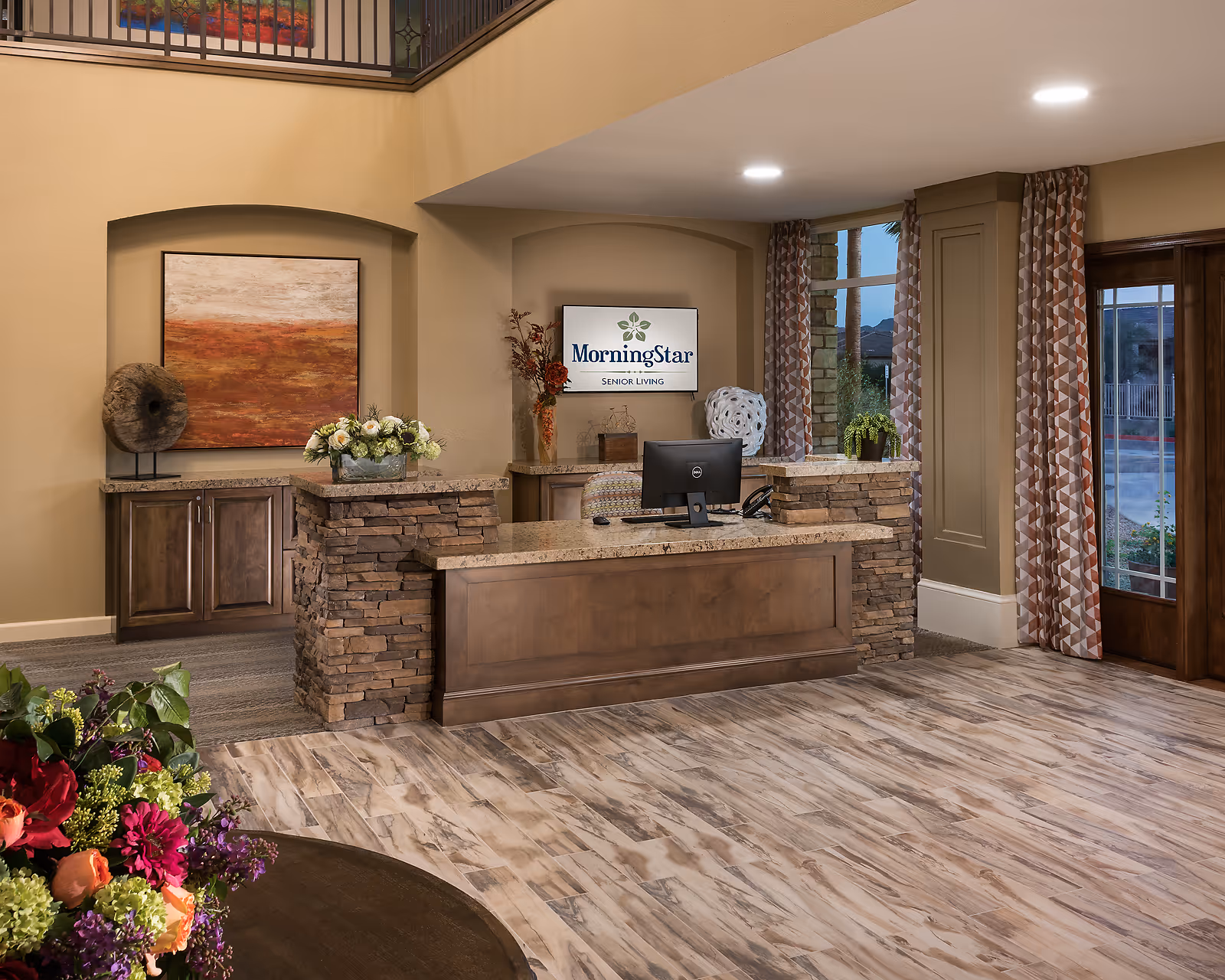 Reception area of MorningStar Assisted Living & Memory Care of Fountain Hills featuring a wooden and stone front desk with a computer, decorative flowers, and artwork on the walls. There are patterned curtains on the windows and a door leading outside.