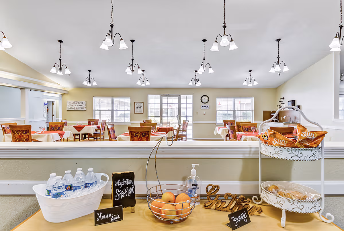 A bright dining room in Meadowview Place with multiple tables covered in white and red tablecloths, surrounded by wooden chairs with red patterned cushions. In the foreground, a hydration station is set up with bottled water in a white tub, a basket of apples, hand sanitizer, and a two-tiered tray holding bags of Sun Chips and pastries. The room has several hanging light fixtures and large windows letting in natural light.