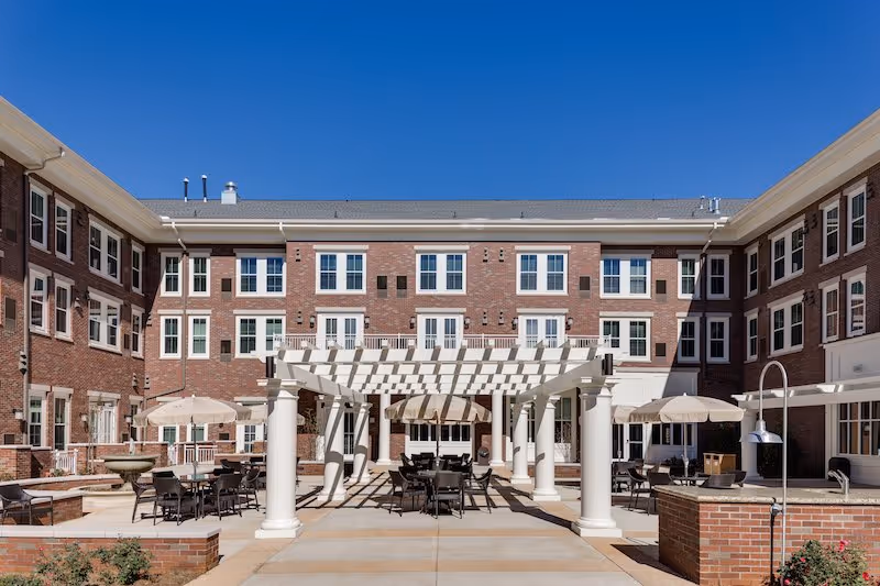 Outdoor courtyard area of a senior living facility with a white pergola, multiple tables and chairs, umbrellas, and a brick building with many windows in the background under a clear blue sky.