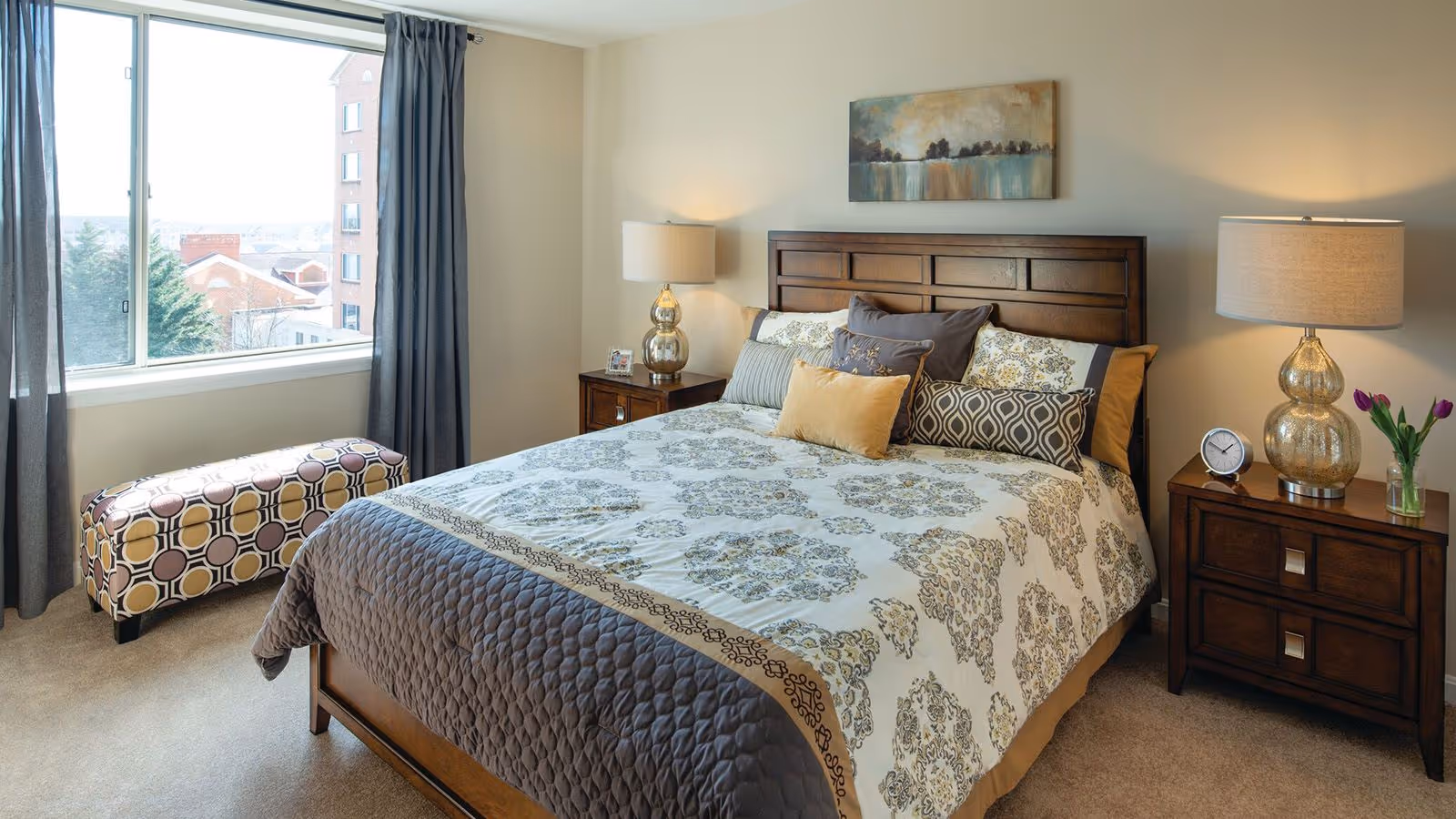 A well-lit bedroom with a large bed featuring patterned bedding and multiple pillows. There are two wooden nightstands on either side of the bed, each with a lamp. A small clock and a vase with purple flowers are on the right nightstand. A window with dark curtains shows a view of buildings and trees outside. A patterned bench is placed under the window.