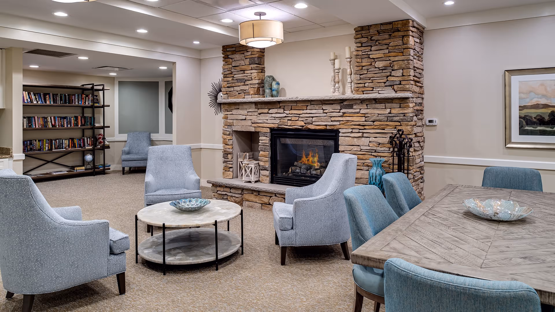 A cozy living room area in a senior living facility featuring a stone fireplace with a fire burning, surrounded by four light blue upholstered armchairs and a round coffee table with a decorative bowl. To the right is a wooden dining table with blue chairs and another decorative bowl. In the background, there is a bookshelf filled with books and additional seating. The room has beige walls, recessed lighting, and a ceiling light fixture above the fireplace.