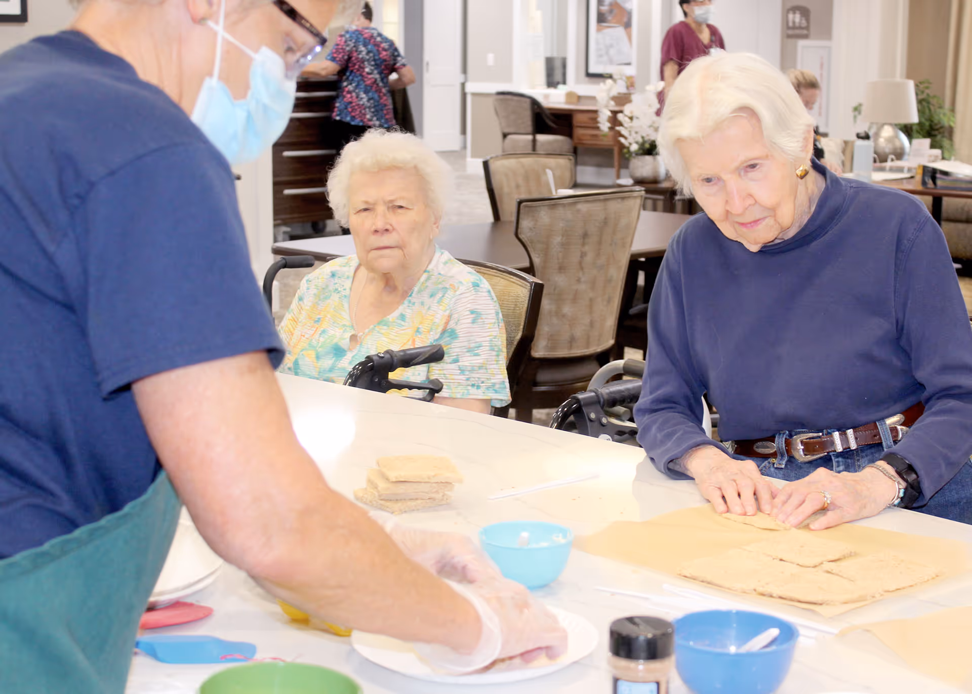 Two elderly women seated at a communal table watching a staff member prepare food in a senior living dining/activity area.