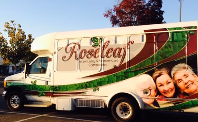 Roseleaf Gardens branded senior living bus parked outdoors beside trees.