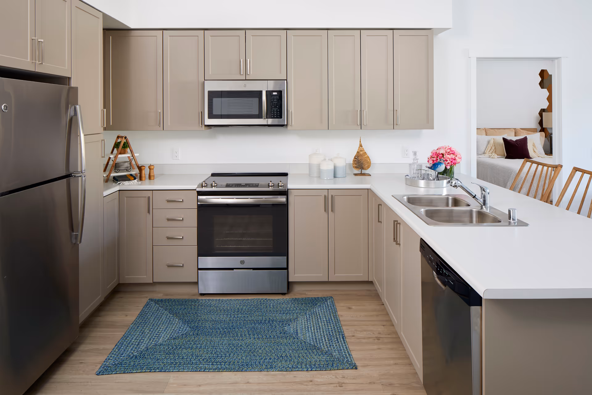Modern open-plan kitchen with beige cabinets, stainless steel appliances, a white countertop island with a double sink, and a blue rug on the wood floor.