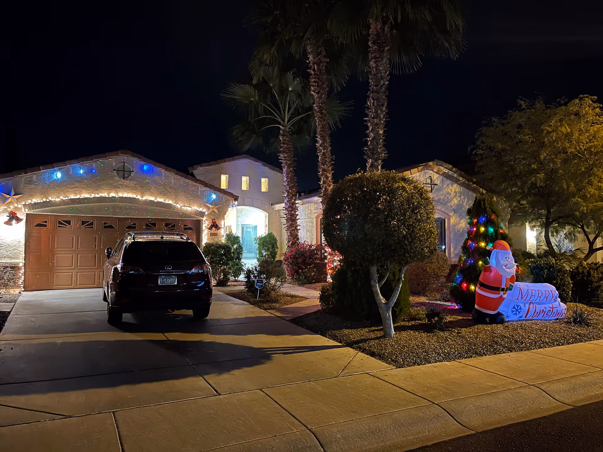 Nighttime view of a residential house decorated with Christmas lights and decorations. The house has a two-car garage with a black SUV parked in the driveway. There are palm trees and trimmed bushes in the front yard, along with an inflatable Santa Claus holding a 'Merry Christmas' sign next to a small Christmas tree with colorful lights.