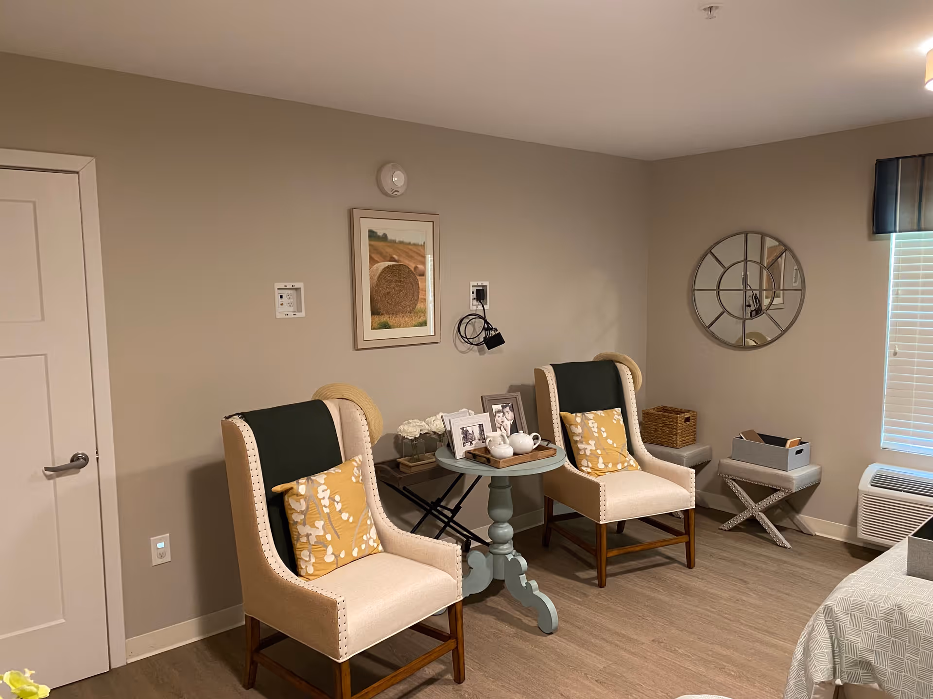 A cozy sitting area in a room with two beige armchairs featuring dark green and yellow patterned cushions. Between the chairs is a small round table with framed photos, a white teapot, and decorative items. The walls are painted light beige with a framed picture of a hay bale and a round decorative mirror. There is a window with blinds and a valance, a small bench with a basket and a box, and a white door on the left side.