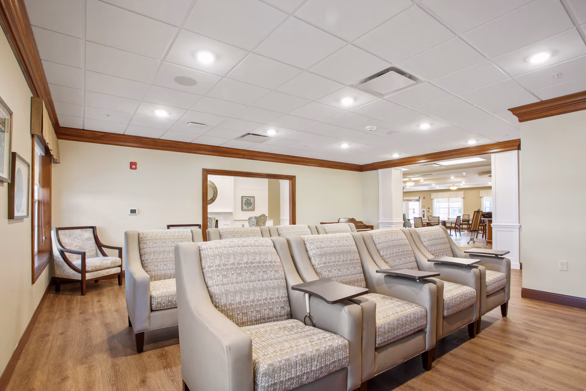 A bright and spacious senior living common area with rows of beige and patterned armchairs equipped with small attached tables. The room features wood flooring, cream-colored walls with wooden trim, recessed ceiling lights, and a large open doorway leading to another seating area with tables and chairs.