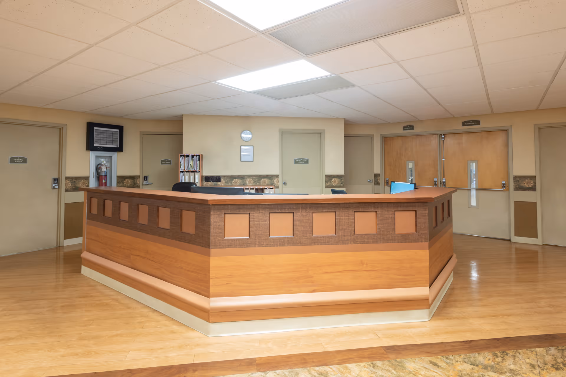 Reception desk area in a health and rehabilitation center with wooden paneling, beige walls, multiple closed doors, a fire extinguisher mounted on the wall, and a ceiling with fluorescent lighting.