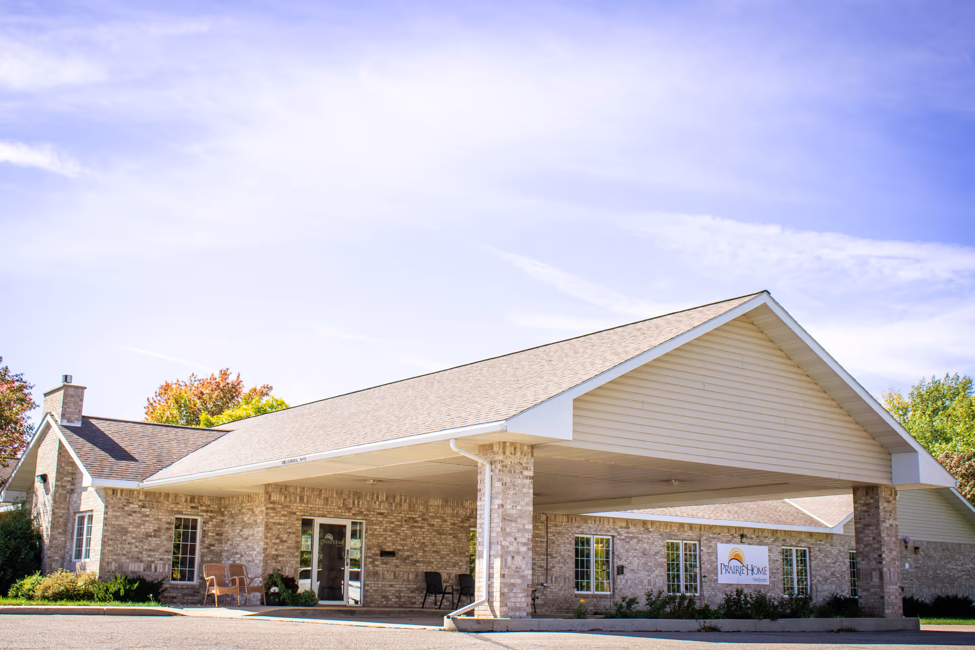 Exterior view of Prairie Home Fieldcrest, a single-story brick building with a large covered entrance and a sign displaying the facility's name. The sky is clear with some clouds, and there are trees with autumn foliage in the background.