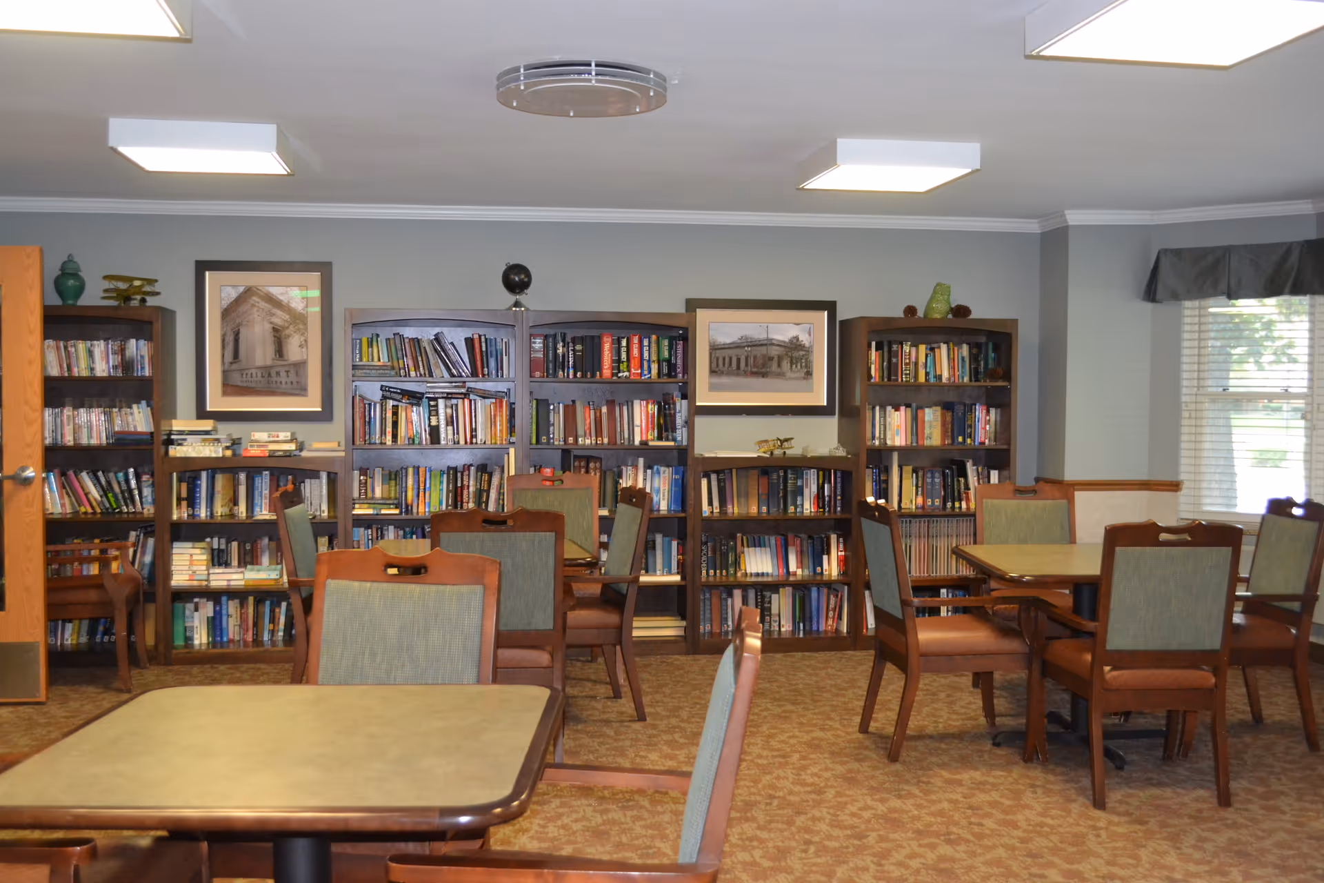 A quiet room with several wooden tables and chairs arranged for seating. The back wall features multiple bookshelves filled with books and framed pictures hanging above them. A window with blinds and a valance is on the right side, allowing natural light into the room.