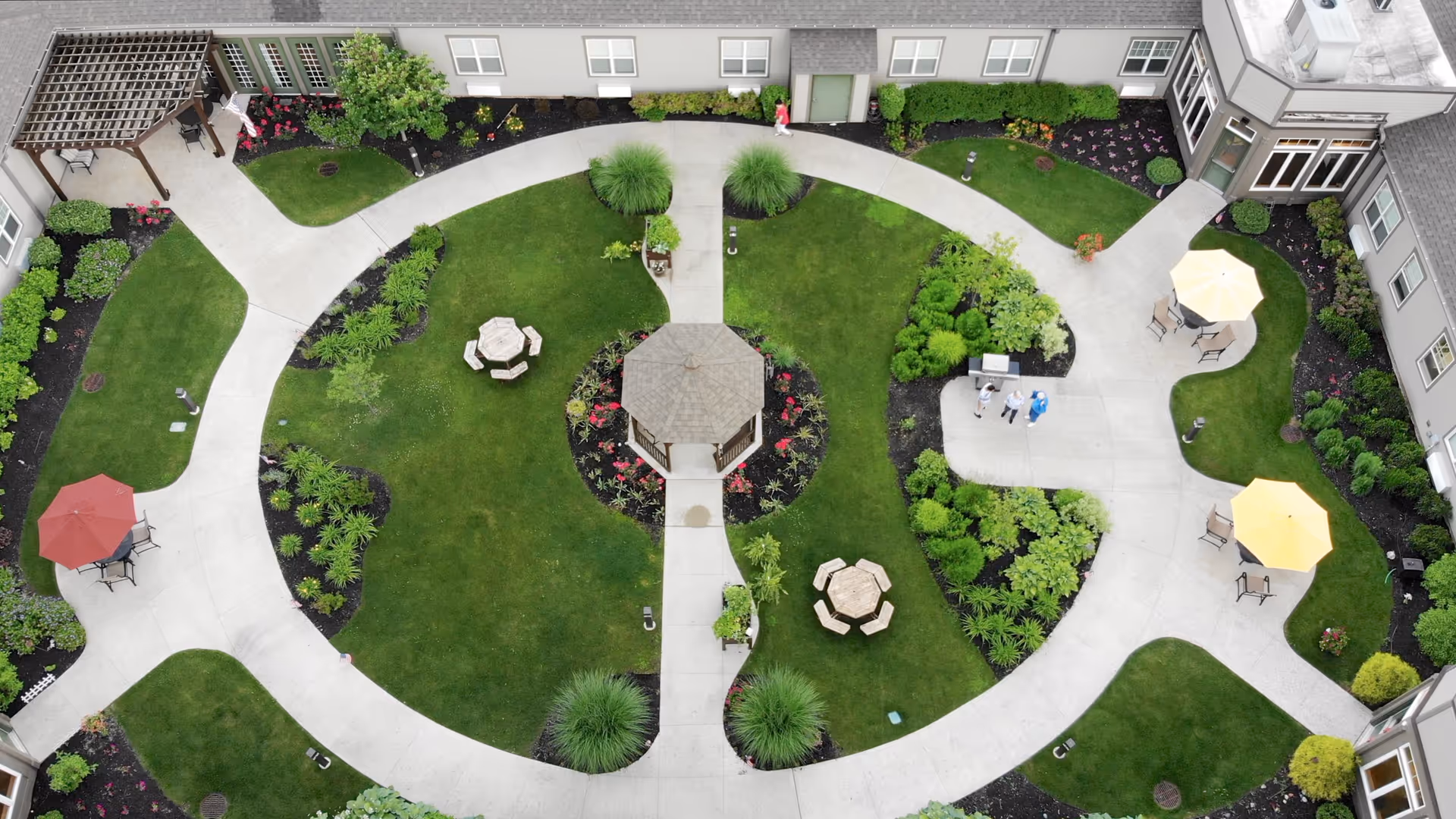 Aerial view of a landscaped courtyard at a senior living facility with green lawns, flower beds, paved walking paths, a central gazebo, picnic tables with umbrellas, and a few people walking.