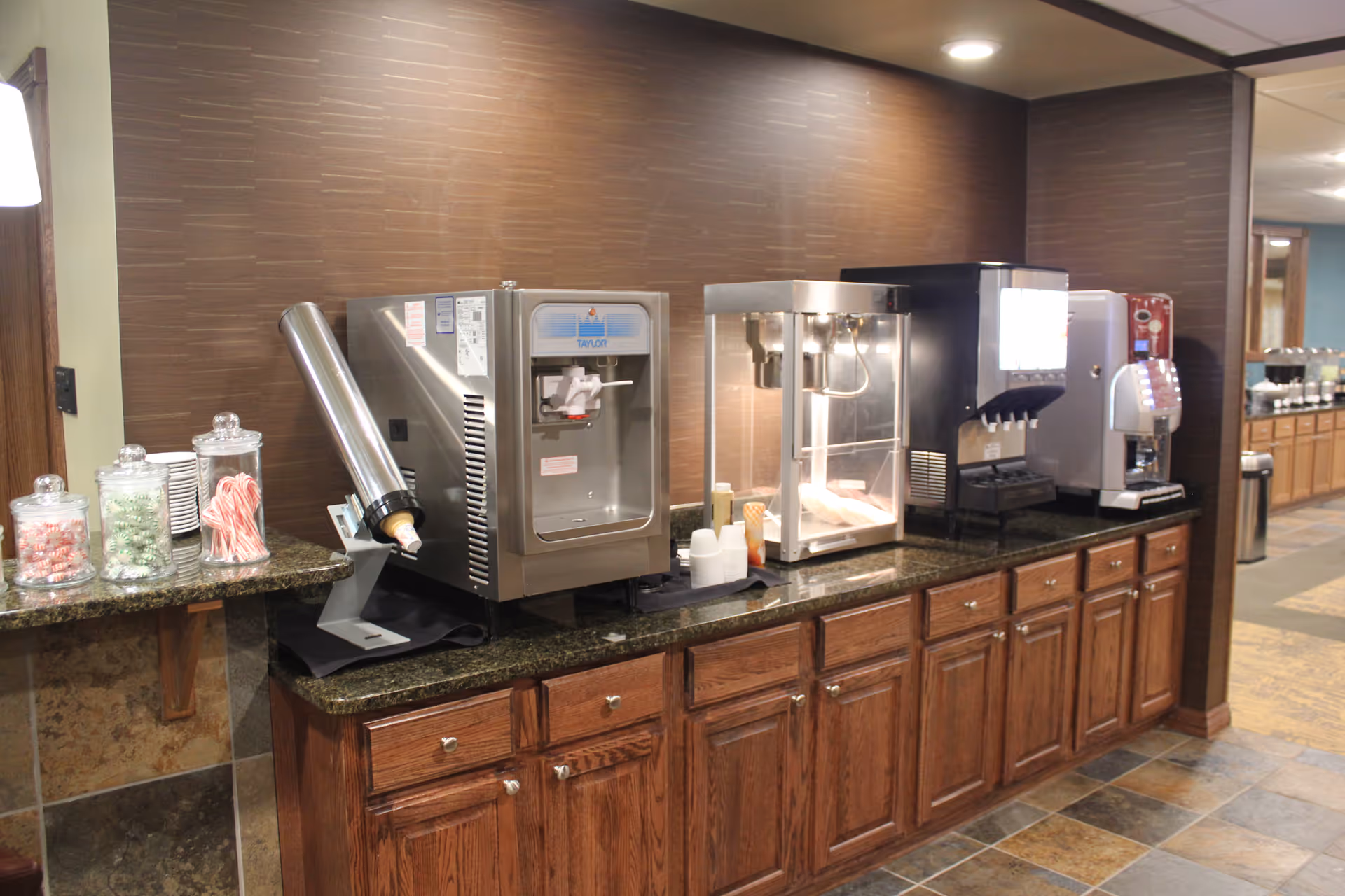 A countertop snack and beverage station with dispensers, a popcorn machine, candy jars, and wooden cabinets in a communal area.