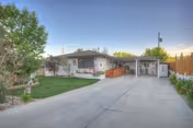Single-story residential building with a concrete driveway, a small lawn with trees and shrubs on the left, and a wooden fence on the right under a clear sky.