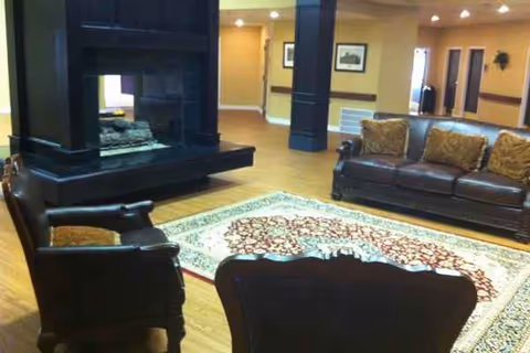 Spacious senior living lounge with leather seating, a fireplace, and a patterned area rug.