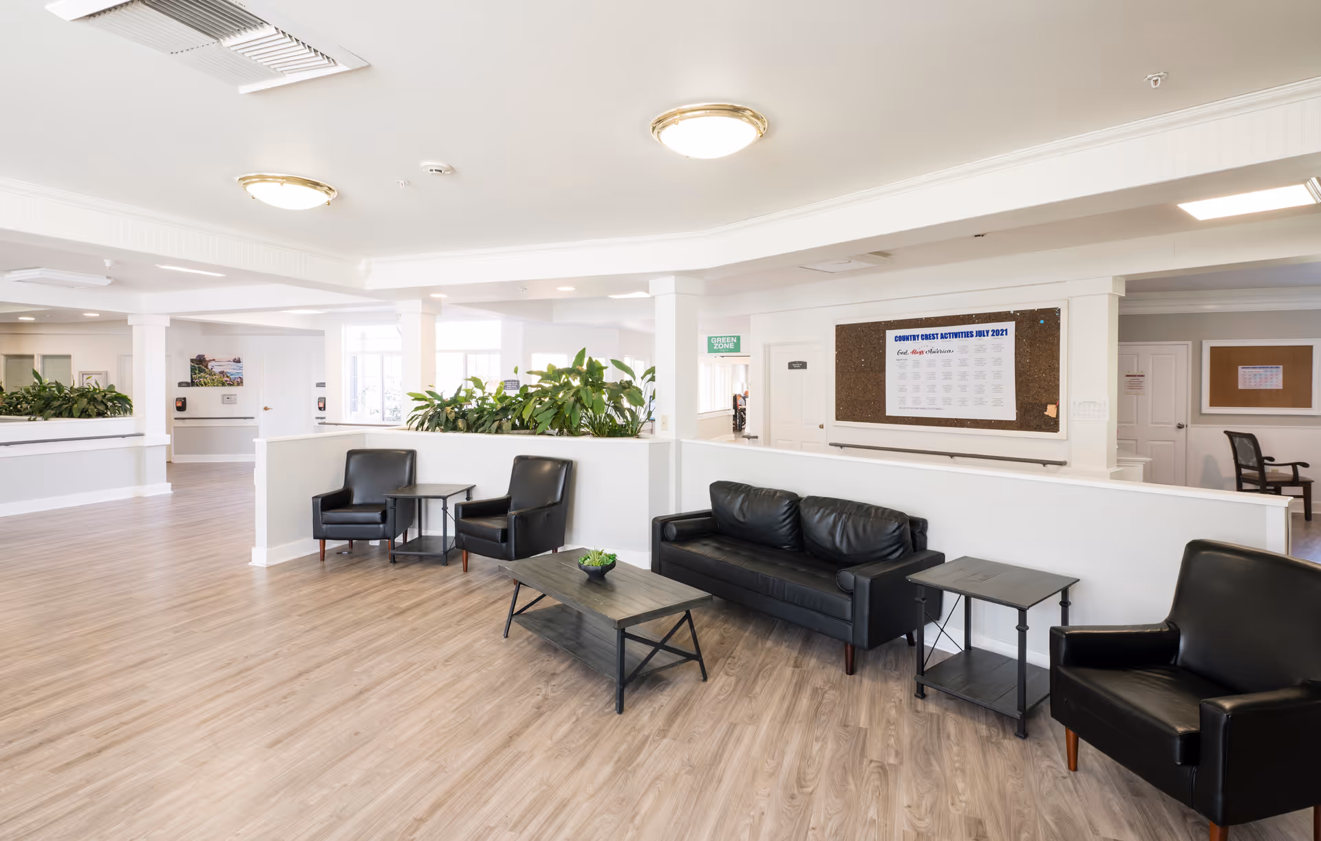 A bright and spacious common area in a senior living facility with light wood flooring, white walls, and ceiling. The room features black leather seating including a sofa and armchairs arranged around a coffee table with a small plant. There are green plants on a partition wall, and bulletin boards with notices on the walls. The space is well-lit with ceiling lights and natural light from windows.