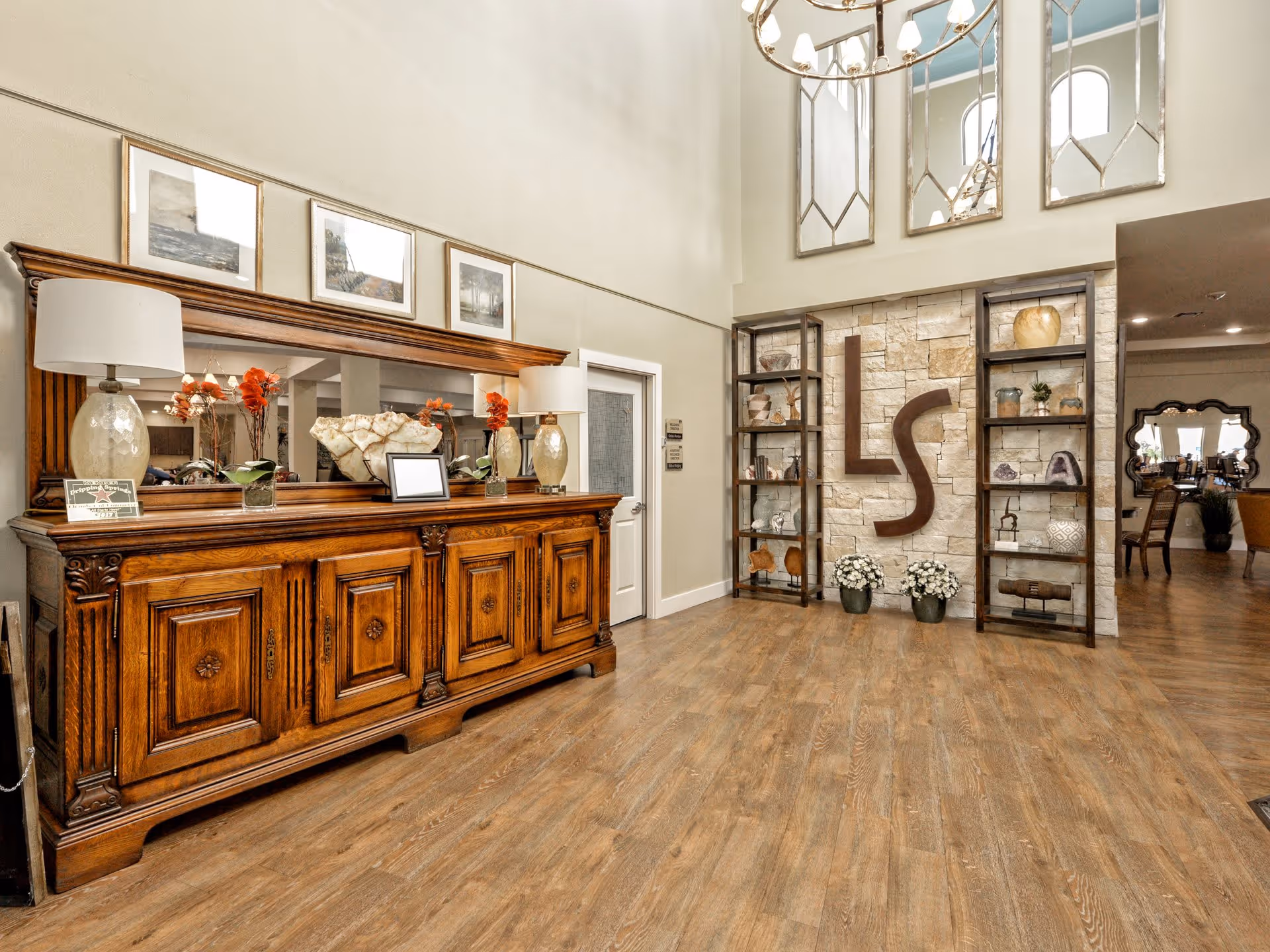 Interior view of a senior living facility lobby area with wooden flooring, a large wooden sideboard with lamps and decorative items, three framed pictures above the sideboard, and a stone wall with large decorative letters 'LS' flanked by two shelving units holding various ornaments. A chandelier hangs from the ceiling, and a doorway leads to another room with more seating visible.