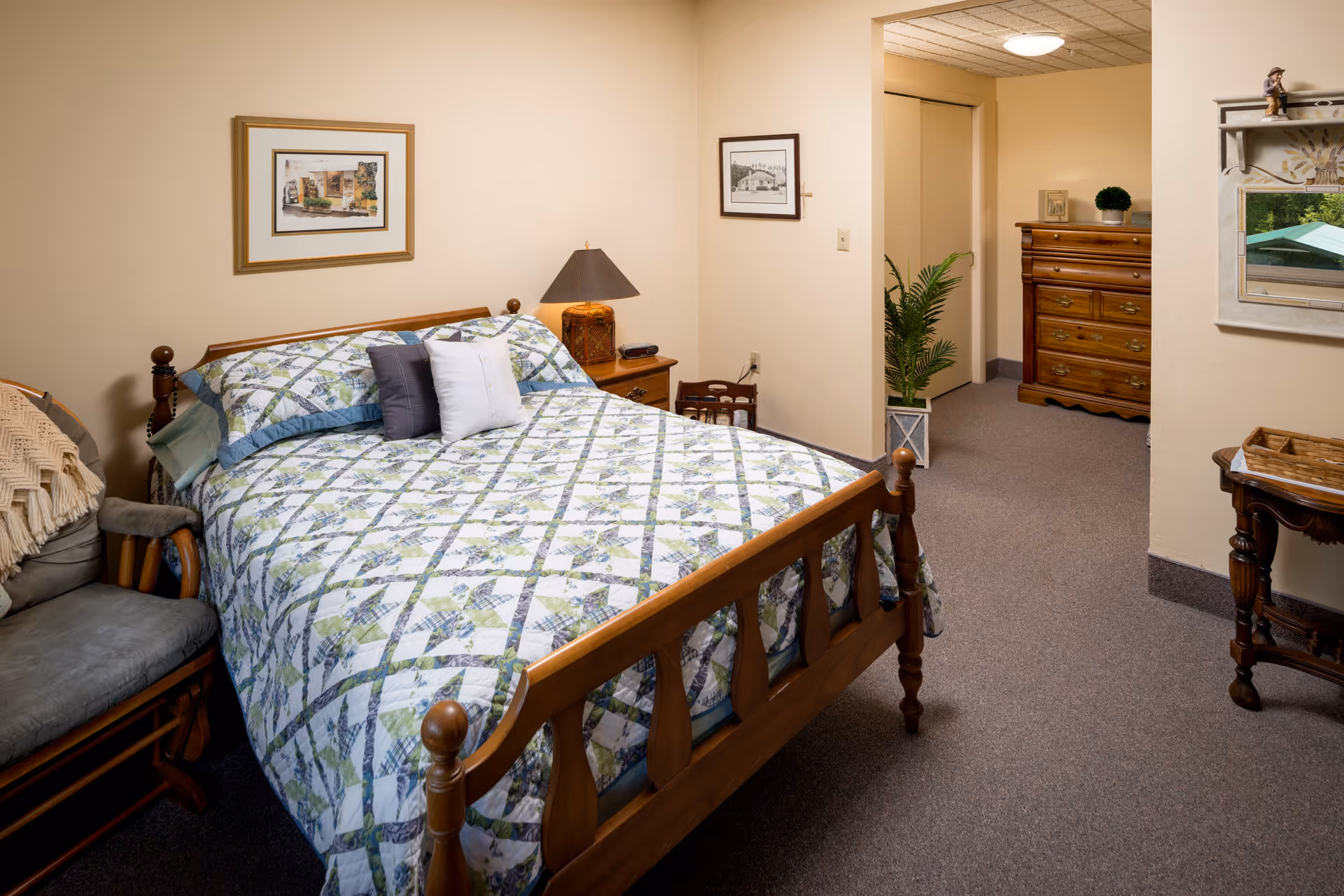 A cozy bedroom with a wooden bed frame and a quilted bedspread in blue, green, and white patterns. There are two pillows on the bed, a wooden nightstand with a lamp, and framed pictures on the beige walls. A cushioned chair with a knitted throw is next to the bed. In the background, there is a wooden dresser, a potted plant, and a small table with a basket. The room has carpeted flooring and soft lighting.