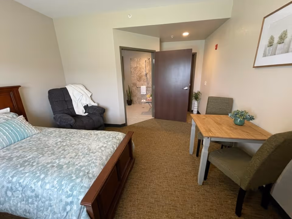 A bedroom in an assisted living facility featuring a bed with patterned bedding, a dark gray recliner chair with a white throw blanket, a wooden table with two green upholstered chairs, and an open door leading to a bathroom with tiled walls and a shower.
