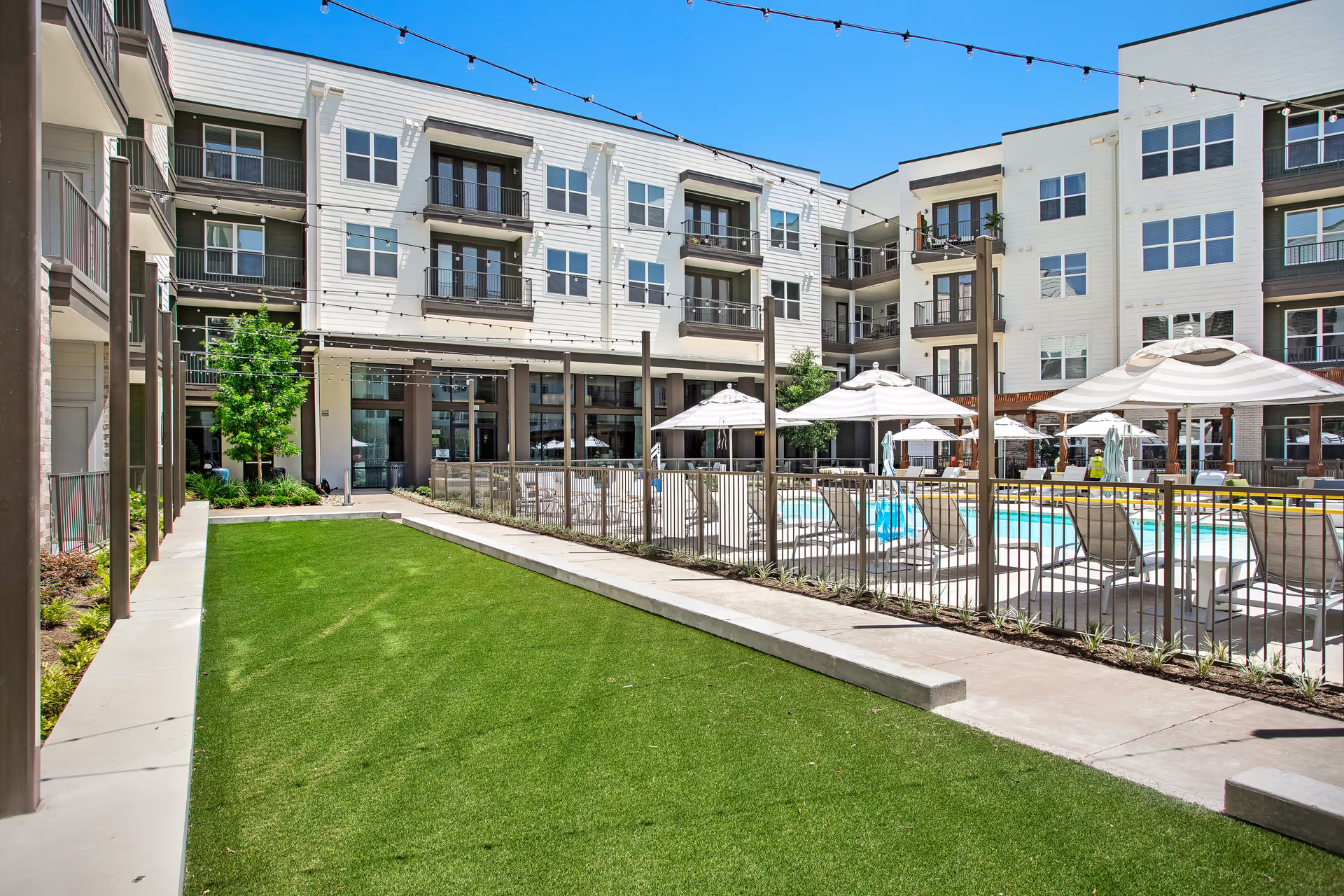 Outdoor courtyard area of a senior living facility with a green lawn, a fenced swimming pool surrounded by lounge chairs and umbrellas, and a multi-story building with balconies in the background under a clear blue sky.