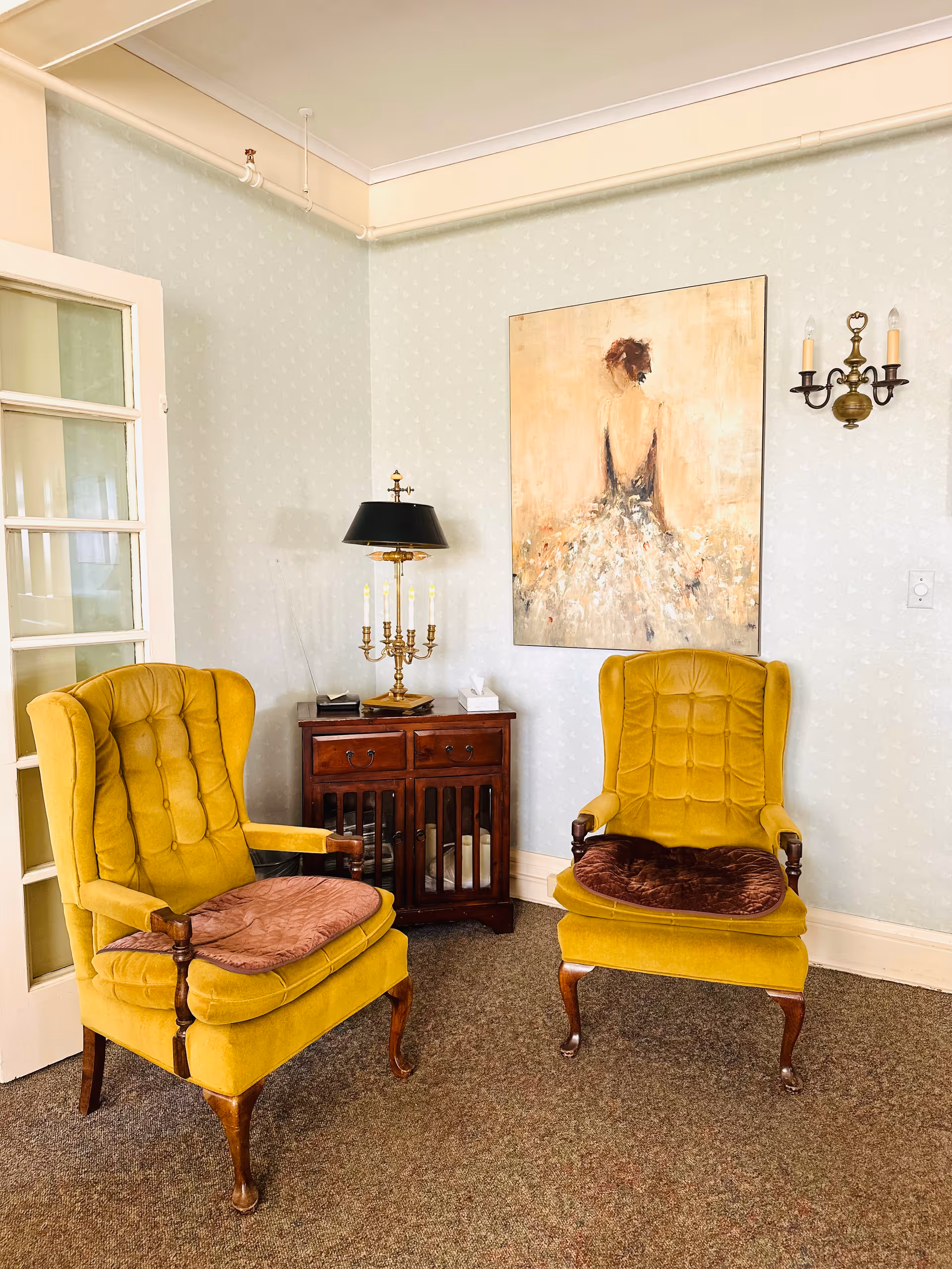 A cozy corner with two vintage yellow upholstered armchairs with brown cushions, a wooden cabinet with a black lamp and a candelabra on top, and a painting of a woman in a dress hanging on a light patterned wall.