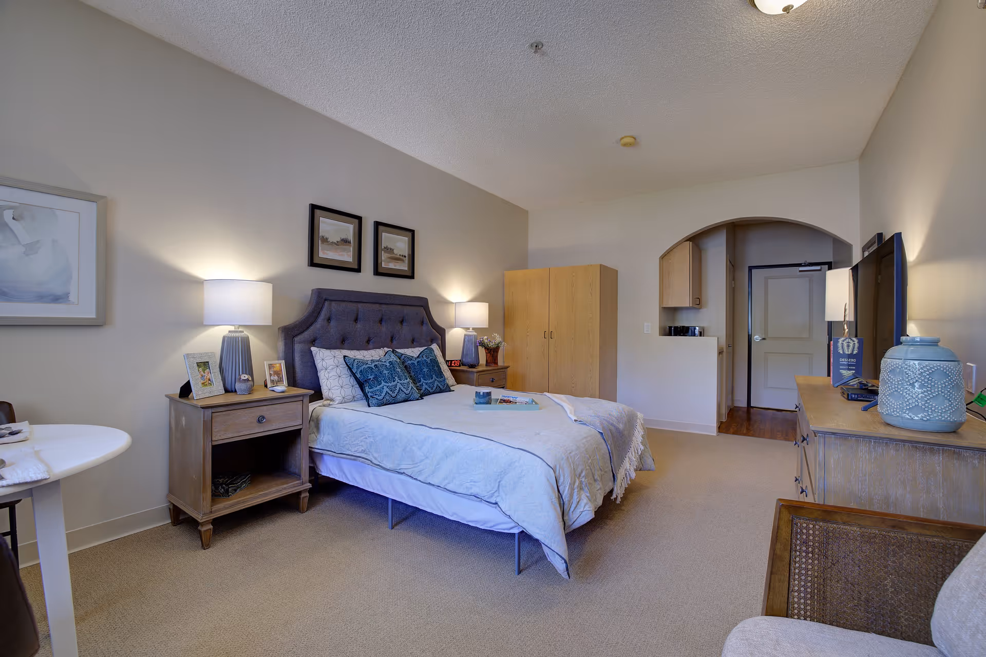 A cozy senior living bedroom with a neatly made bed featuring blue and white bedding and decorative pillows. Two bedside tables with lamps and framed photos flank the bed. On the right side, there is a wooden dresser with a large TV and decorative items. The room has beige walls, carpeted floor, and an arched doorway leading to another area. A small round table and chair are partially visible on the left.