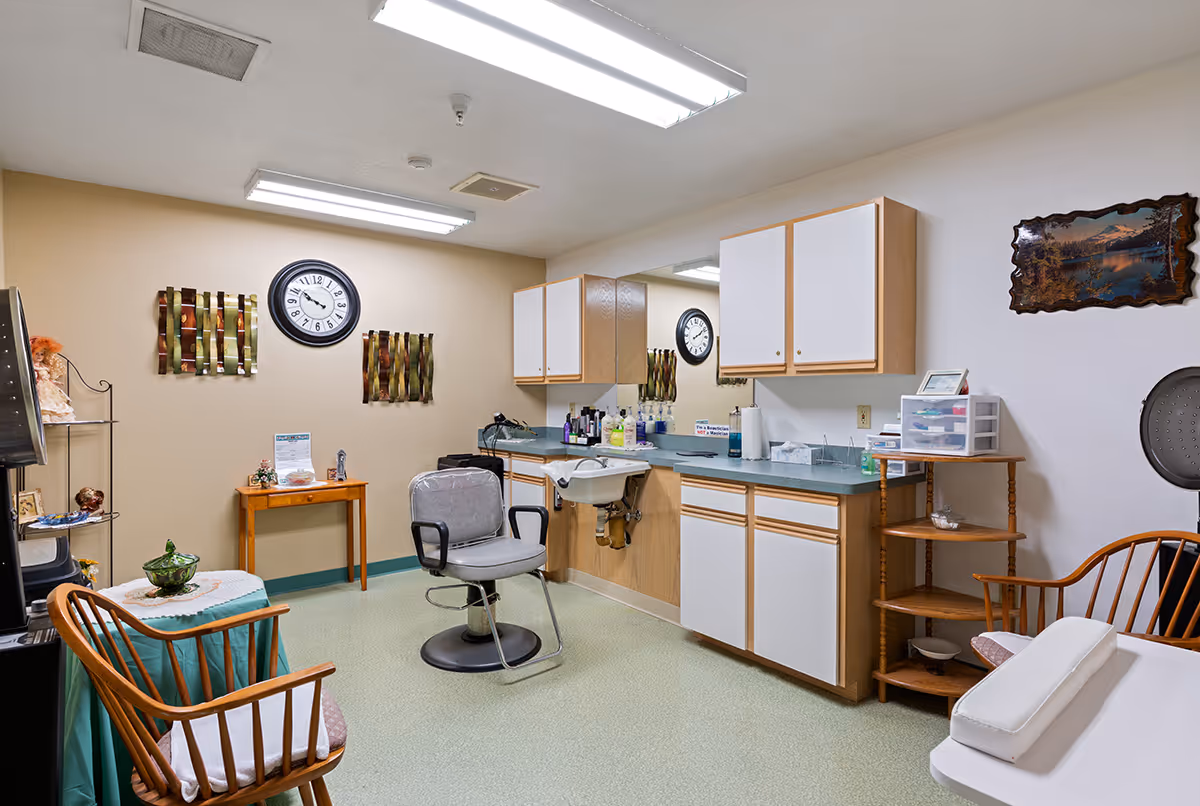 A small salon or grooming room with a salon chair in front of a sink and counter with various hair and beauty products. The room has light-colored walls with a clock and decorative wall hangings. There are wooden chairs with cushions, a small table with a green cloth, and cabinets mounted on the wall. The floor is covered with light green linoleum, and fluorescent lights illuminate the room.