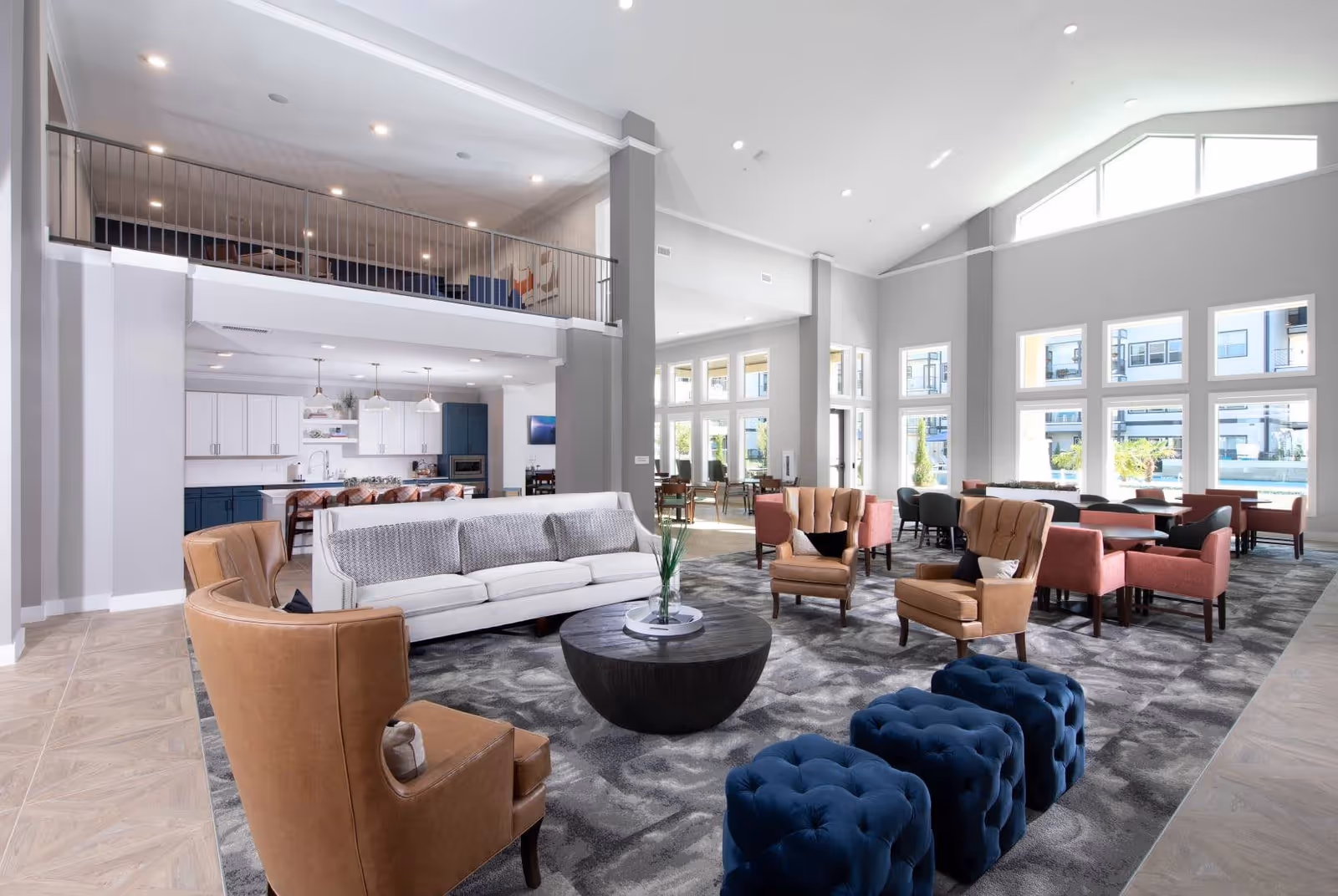 A spacious, modern common area with high ceilings and large windows letting in natural light. The room features a white sofa, brown armchairs, blue tufted ottomans, and several tables with chairs arranged for socializing or dining. In the background, there is a kitchen area with white and blue cabinetry and pendant lighting.