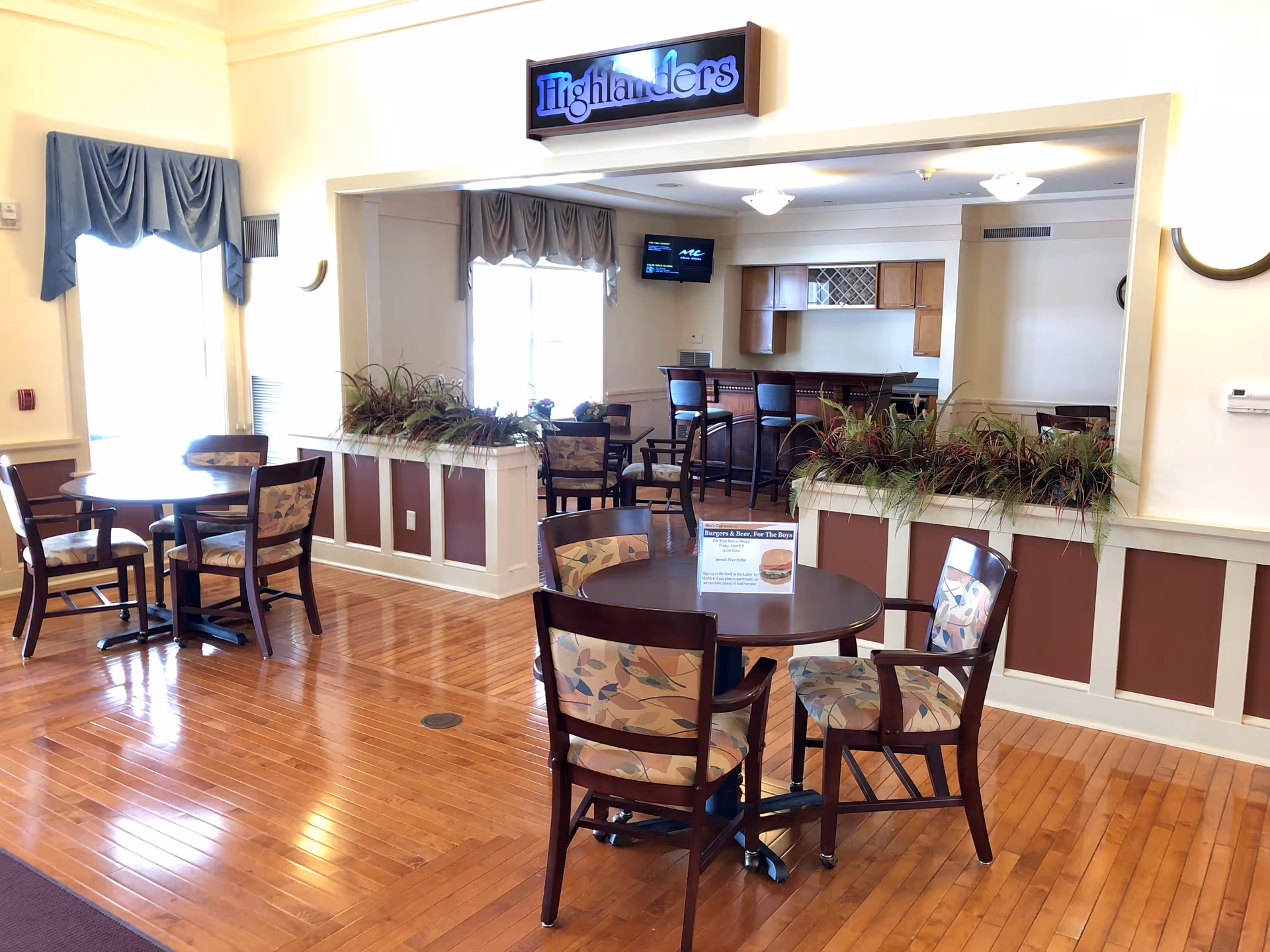 A dining area with wooden floors and several round tables surrounded by chairs with patterned cushions. There is a counter with bar stools in the background and a sign above that reads 'Highlanders'. The room is well-lit with natural light coming through windows with blue curtains.