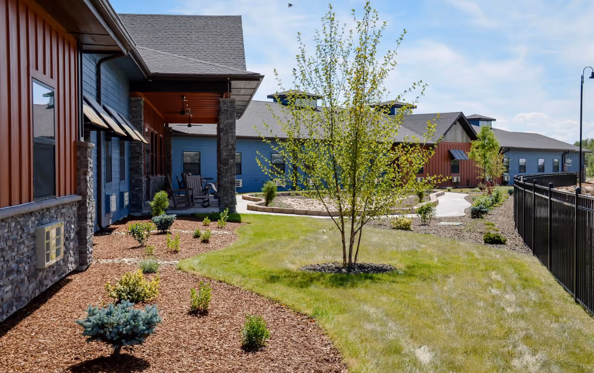 Outdoor view of Aspen Valley Senior Living facility showing a landscaped garden area with small trees, shrubs, and a grassy lawn. The building has a combination of stone and wood siding with a covered porch featuring rocking chairs. A paved walkway curves through the garden, and a black metal fence runs along the right side.