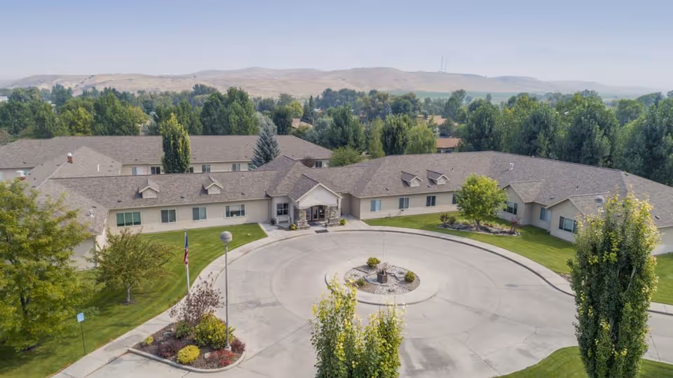 Aerial view of a single-story senior living facility building with a circular driveway and landscaped greenery including trees and bushes. The building is surrounded by trees and hills in the background under a clear sky.