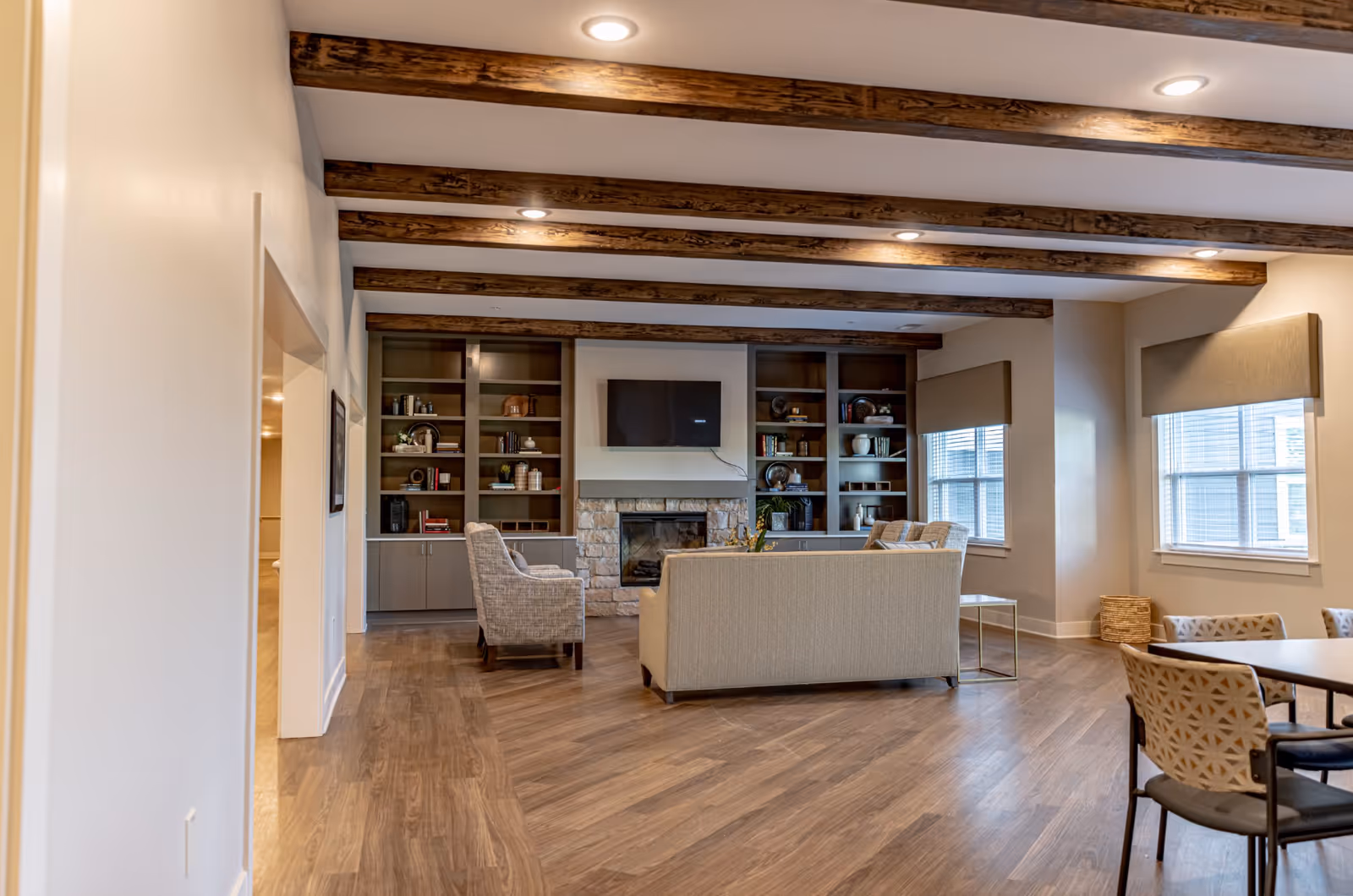 A spacious living room with wooden floor and exposed wooden ceiling beams. The room features a stone fireplace with a flat-screen TV mounted above it, flanked by built-in shelves filled with decorative items and books. There are two armchairs and a sofa arranged around a coffee table, and a dining table with chairs is visible on the right side near windows with beige blinds.