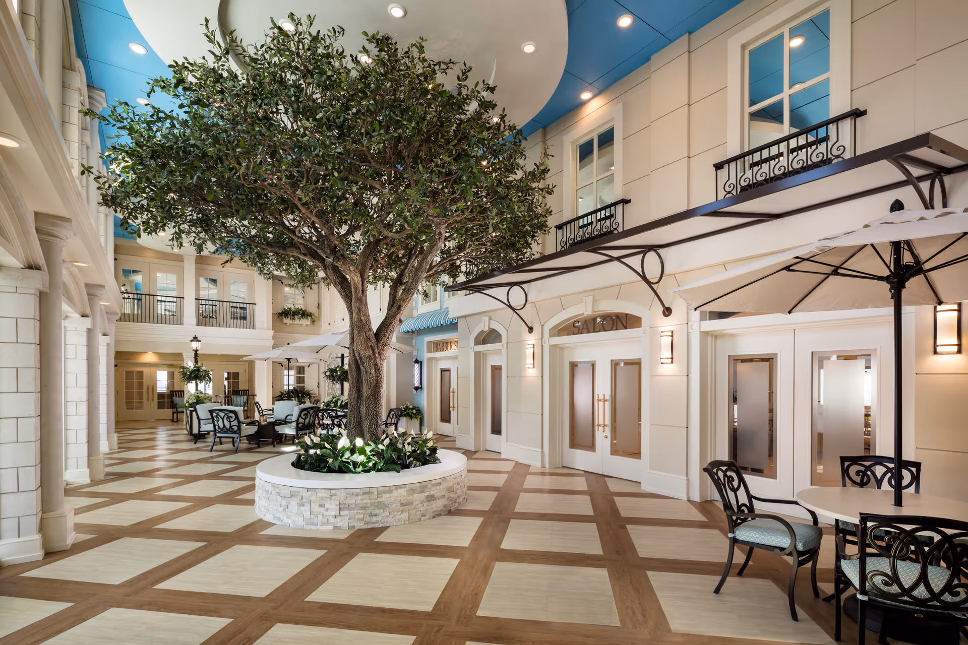 Indoor courtyard area with a large tree planted in a circular stone planter in the center. Surrounding the tree are tables and chairs with umbrellas. The walls have multiple doors and windows, including a door labeled 'SALON'. The ceiling is painted blue with recessed lighting, and the floor has a checkered pattern of wood and light tiles.
