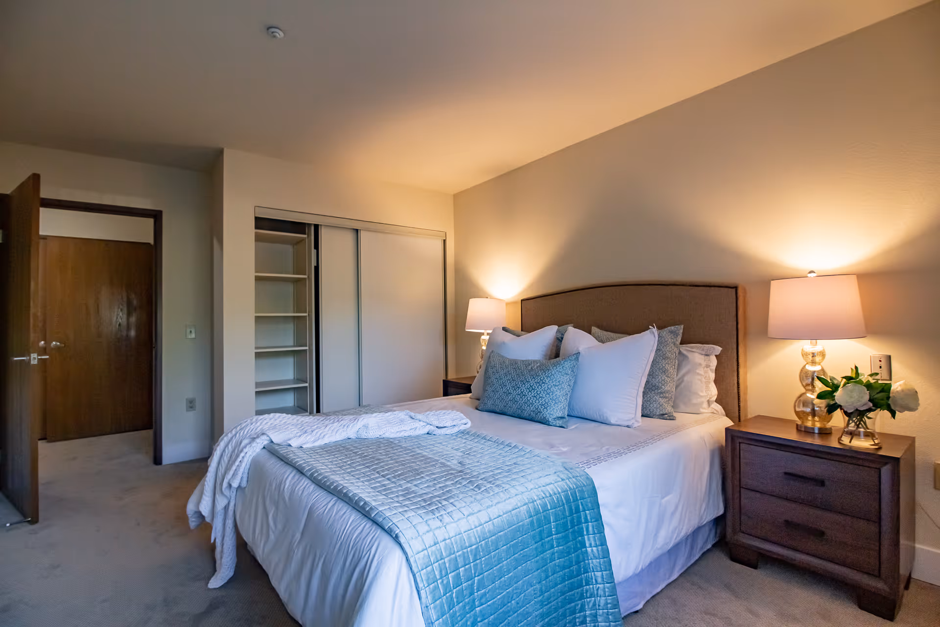 A neatly made bed with multiple pillows and a light blue quilted blanket in a bedroom. There are two bedside tables with lamps and a small vase of flowers on the right table. The room has beige walls, a carpeted floor, an open closet with sliding doors, and a wooden door leading out of the room.