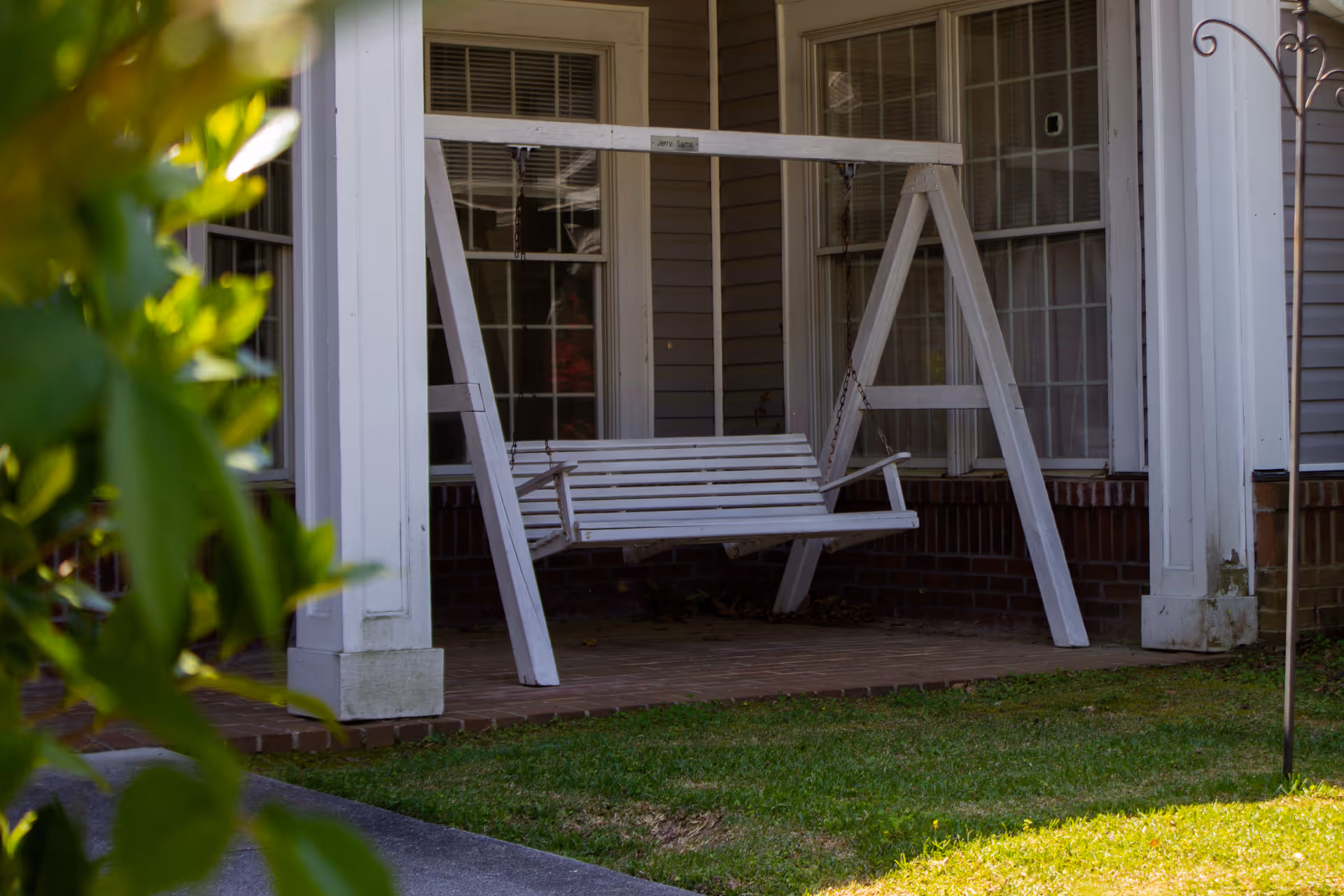 White wooden porch swing hanging from a wooden frame on a covered front porch with windows and greenery in the foreground.