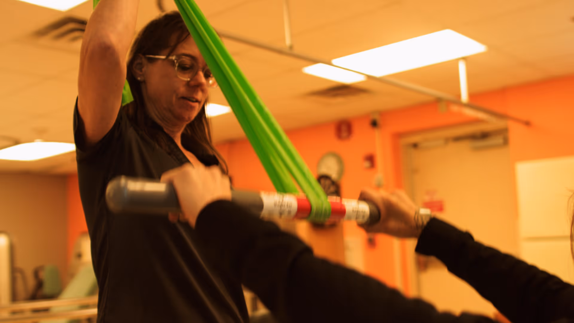 A woman wearing glasses assists another person with a physical therapy exercise using a green resistance band and a gray bar in an indoor room with orange walls and ceiling lights.
