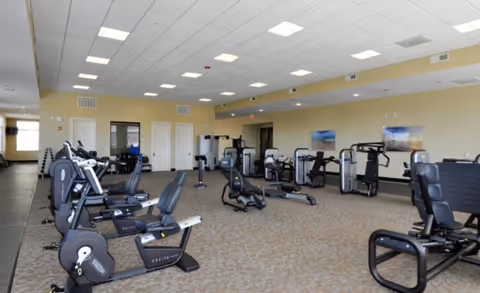 Spacious fitness room with multiple cardio and strength machines arranged on a carpeted floor under a drop ceiling with recessed lights.