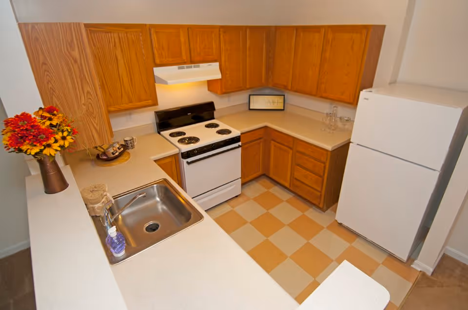 Small kitchen with wooden cabinets, a white stove and refrigerator, a stainless sink, and a checkered floor.