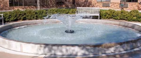 A circular outdoor fountain with water spraying overlooks a brick building and a white bench.