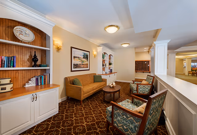 A cozy senior living facility common area with a brown couch, two patterned armchairs, a round wooden coffee table, built-in white bookshelves with books and decorative items, wall sconces, and a framed landscape painting on the wall. The carpet has a geometric pattern and the space is softly lit with ceiling lights.