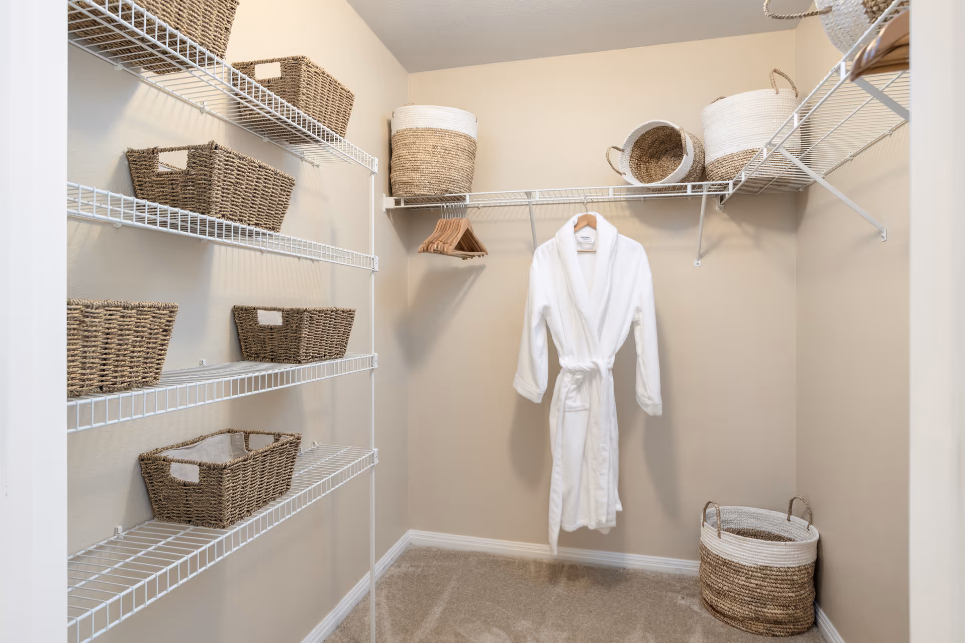 A walk-in closet with beige walls and carpeted floor. The closet has white wire shelving on the left and back walls, holding several woven baskets. A white bathrobe is hanging on a wooden hanger from the back wall shelf, along with additional empty wooden hangers. There are also larger woven baskets placed on the floor and top shelves.