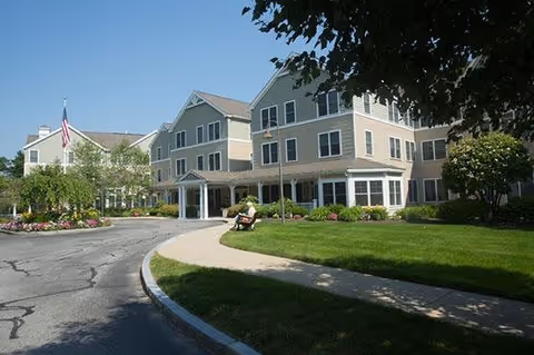 Exterior view of a multi-story senior living facility with beige siding and white trim. There is a curved driveway leading to the entrance, a person sitting on a bench near the sidewalk, well-maintained green lawns, flower beds, and trees providing shade.